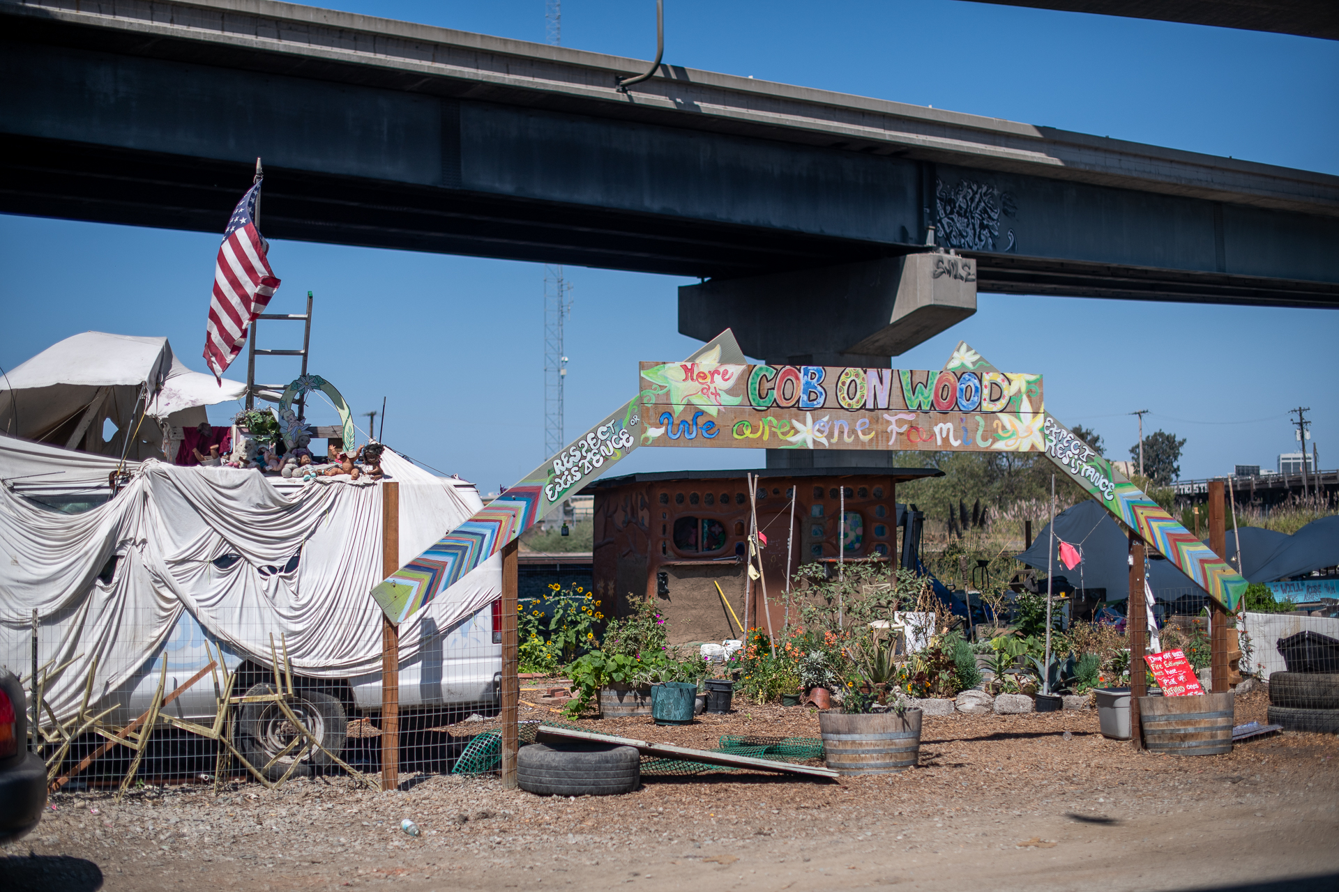 A small, tan shack under the freeway with a garden in front of it and a rainbow sign above a wooden archway is hand painted and reads, "Cob on Wood."