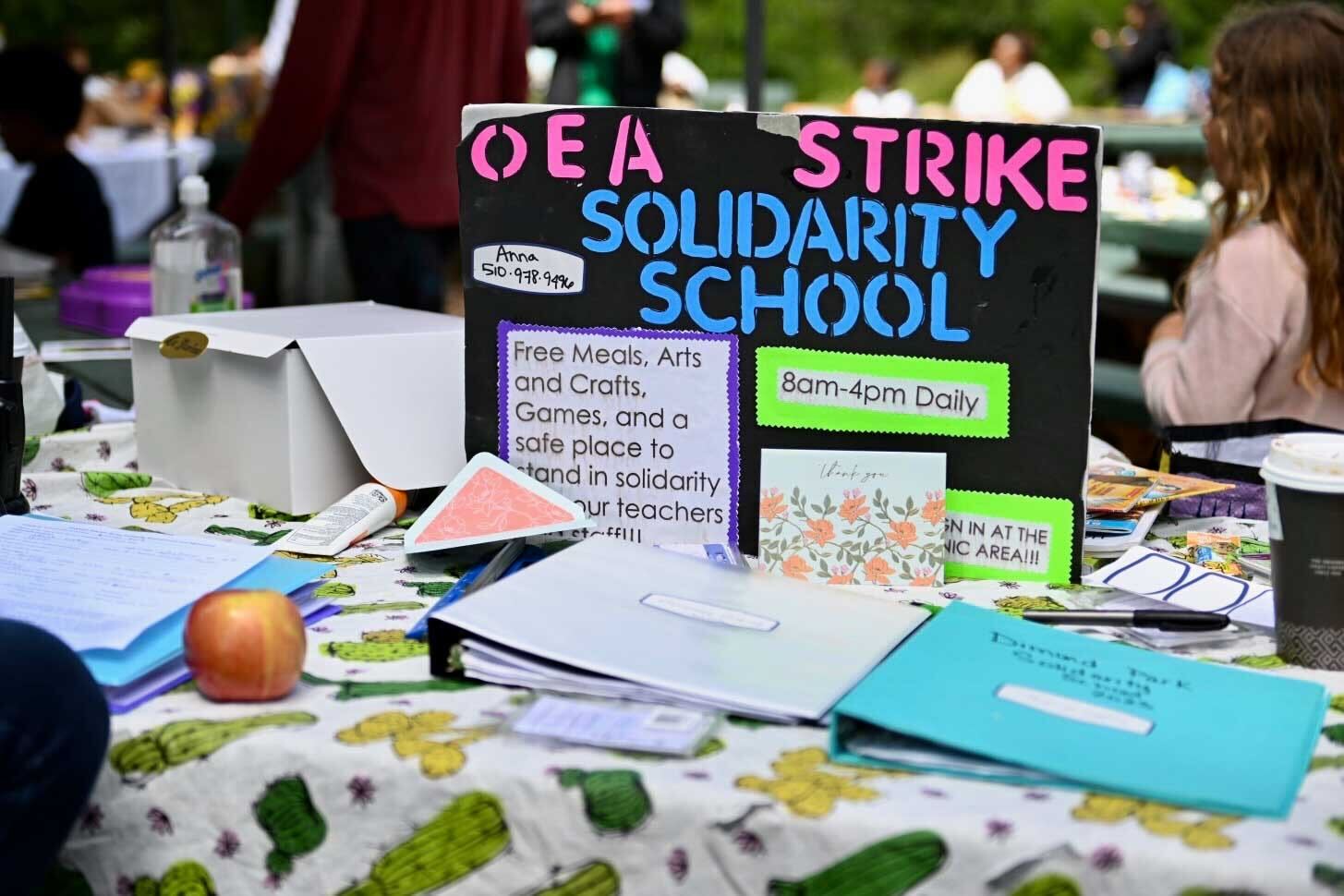A black sign with pink and blue writing sits on a table at a park. The sign reads, "OEA Strike Solidarity School." It also reads, "Free meals, arts and crafts, and a safe place to stand in solidarity with our teachers and staff!" There is a single apple on the table, along with blue and white, three-ring binders. A white pastry box and a bottle of hand sanitizer are also on the table. Parents and children are blurry in the background.