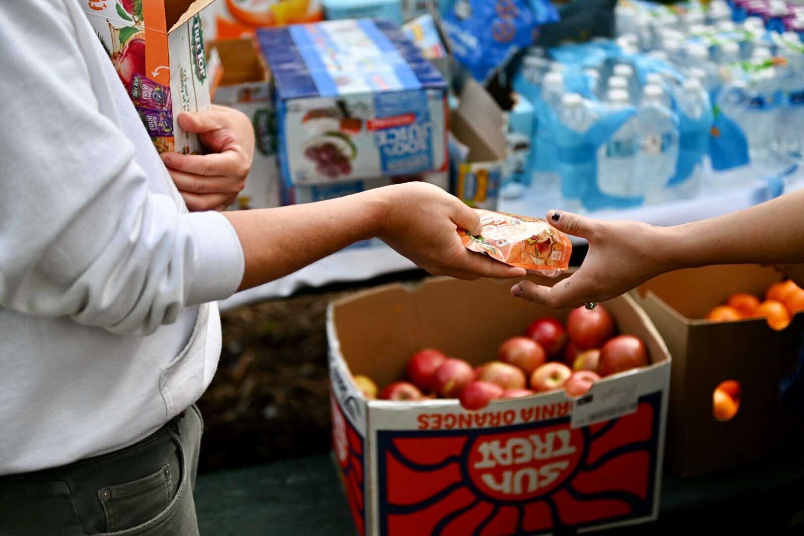 A close-up shot of a grown-up's hand, handing a wrapped snack to a child's hand. A box of red apples and a box of tangerines, along with pallets of water bottles and juice boxes are pictured in the background placed on a park picnic table.