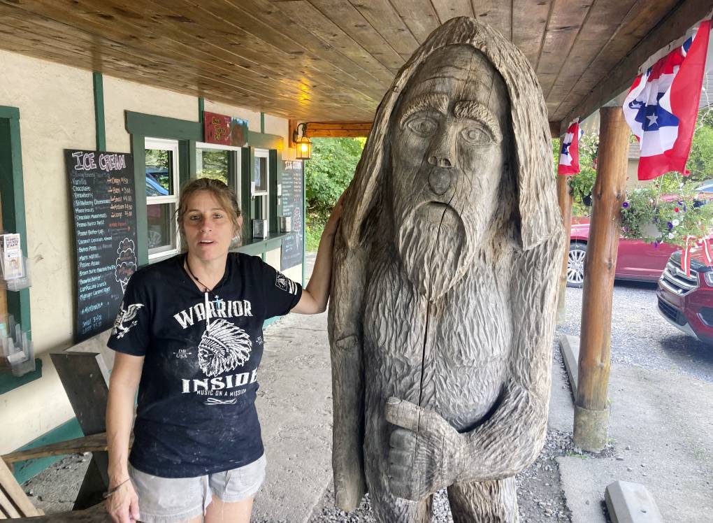 A white woman wearing a black t-shirt that says "Warrior" with a logo and shorts leans her left arm against a wooden statue outside a building.
