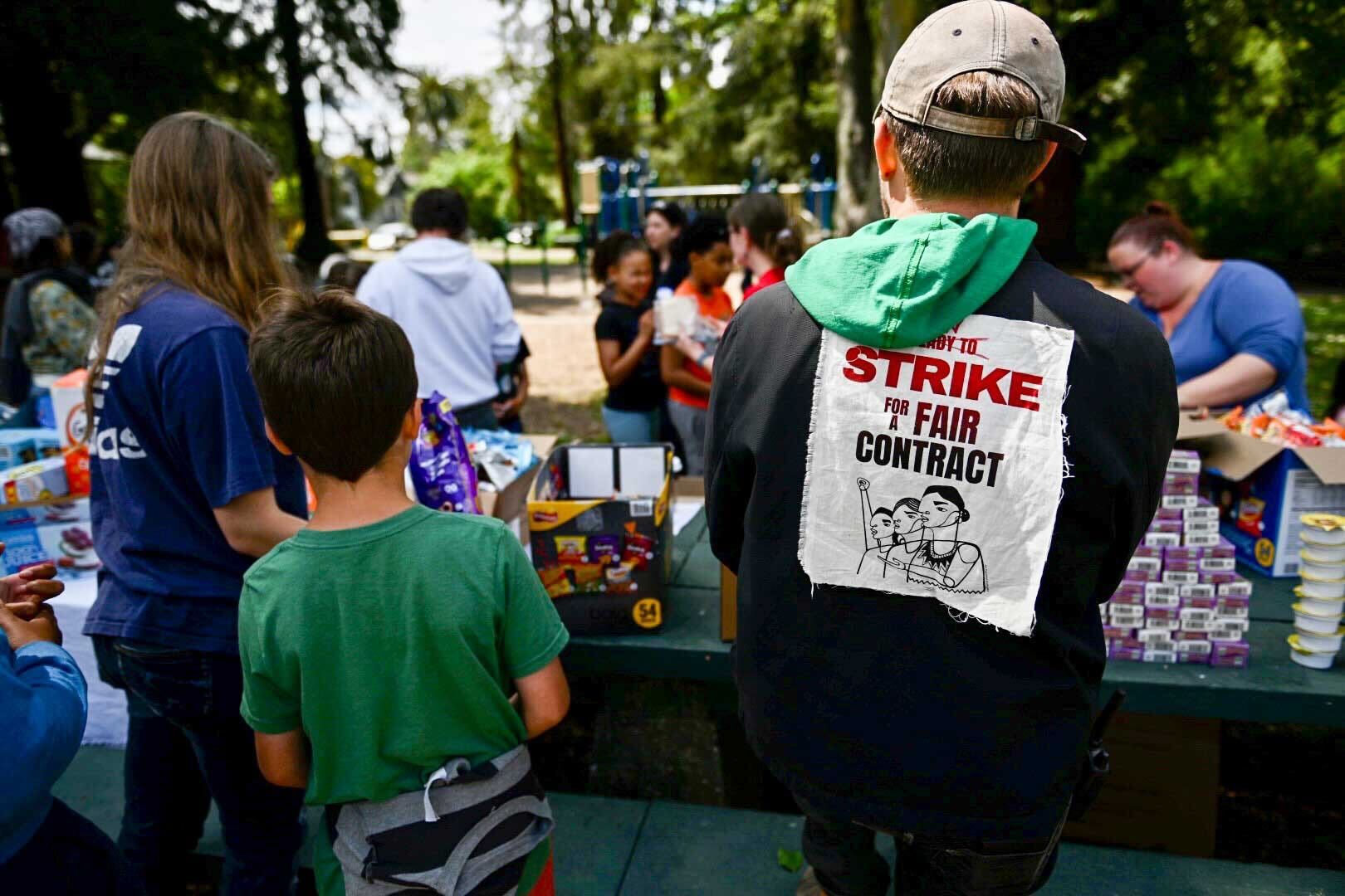 A man wears a black jacket with a white patch with red and black print that reads,"Strike for a fair contract." He stands next to a boy wearing a green T-shirt. The two are in front of a green picnic table that has many snacks and juice boxes on top of it. Many children and parents are seen in the background, along with a blue playground structure shaded by lush trees.