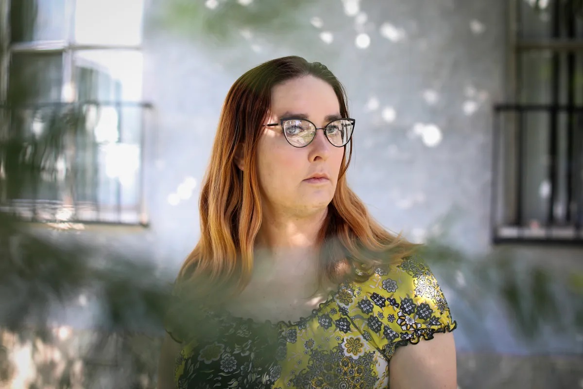 A woman with reddish, shoulder-length hair, cateye glasses and a yellow and black floral blouse poses with a serious face in front of her apartment complex.