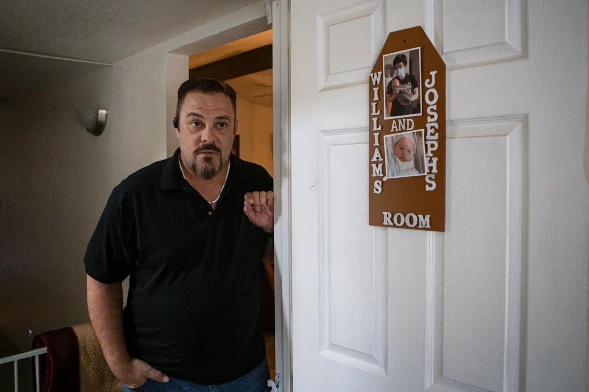 A man wearing a black shirt, gold chain and a black Bluetooth device in his ear poses inside his home next to a white door. On the white door is a homemade sign that reads, "William's and Joseph's Room" with two photos of the two boys.