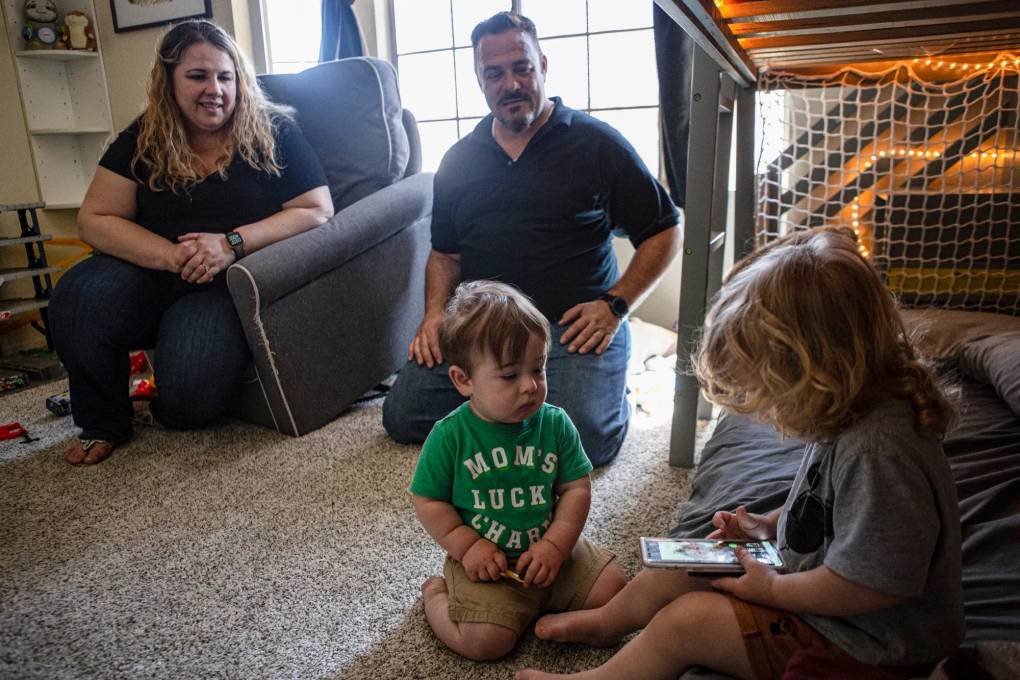 A mother sits in a cushioned, chair inside a child's nursery in her home. She watches her two toddlers play on the tan carpet. Her husband is on his knees watching their children. One child's green T-shirt reads, "Mom's Lucky Charm" in white letters.