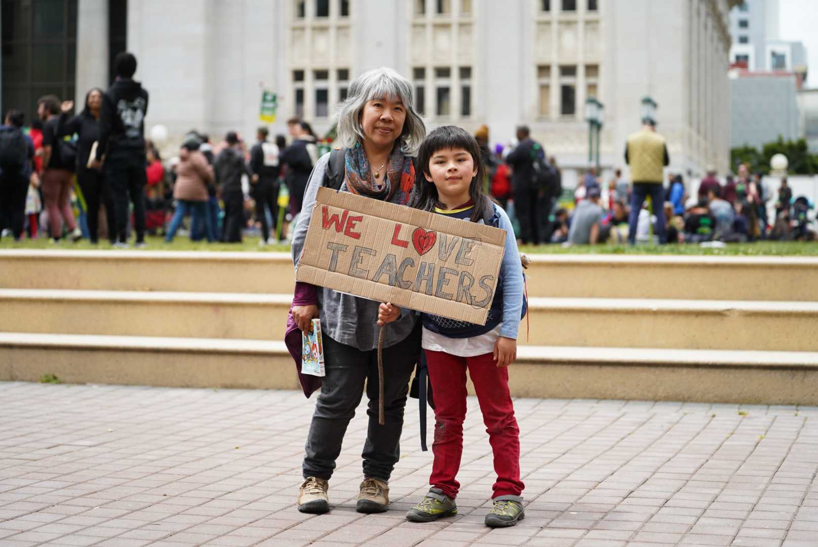 Misa Takaki and her son, Akira Takaki, hold a sign that reads "We Love Teachers" at Thursday's noontime rally at Frank H. Ogawa Plaza. 