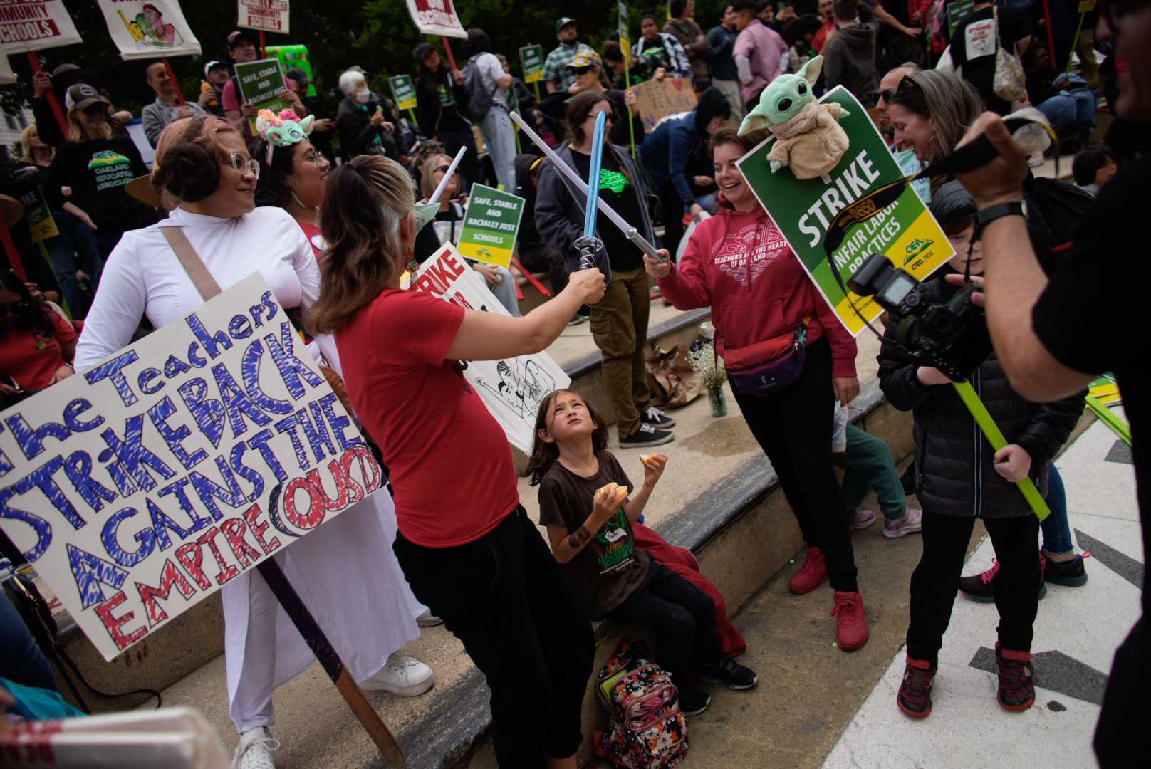 Protestors gather and hold signs, with two female protestors engaged in a mock sword fight.