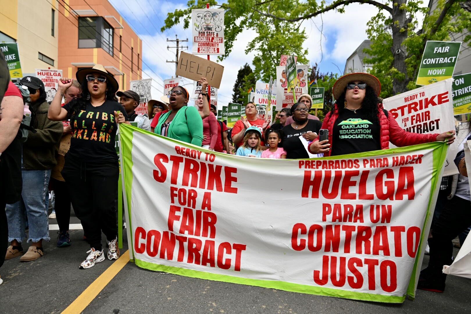 a group of people which appears to be multi-ethnic in composition walks down a street carrying a large banner reading 'ready to strike for a fair contract' in both English and Spanish
