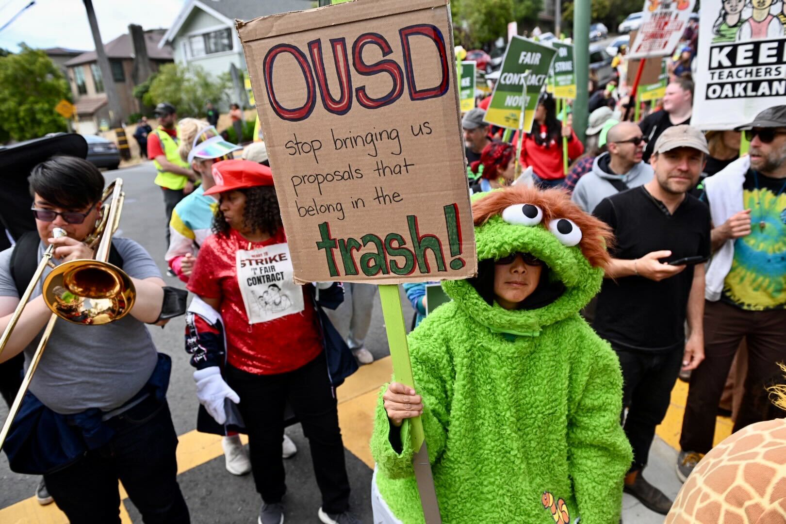 In the foreground is what appears to be a middle school student in a full-body, bright green Oscar the Grouch costume, with a fuzzy brown unibrow, big googly eyes, and the person's face inside the mouth, holding a cardboard sign on a flat wooden stick that says, 'OUSD, Stop bringing us proposals that belong in the trash!' Oscar is in a crowd of people in a street, including someone to their right playing a trombone.