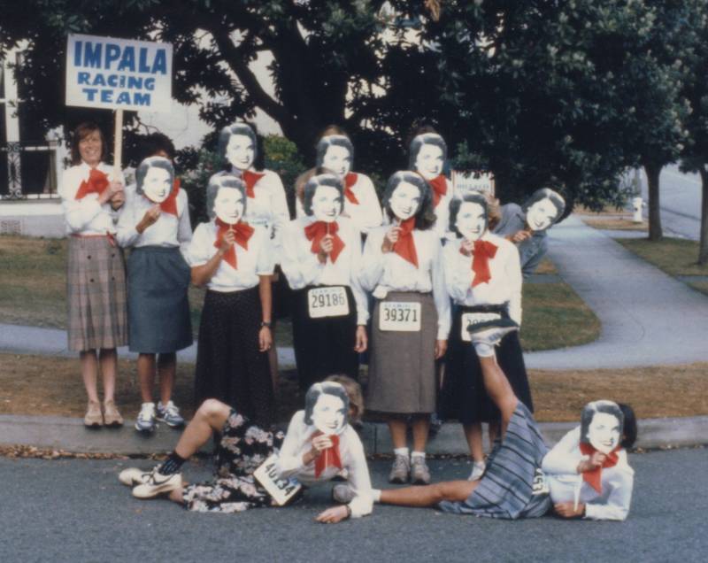 A dozen women wear knee length business skits, white blouses, and identical large red bowties. They are each holding a paper cutout of Dianne Feinstein's face in front of their own, like a mask.
