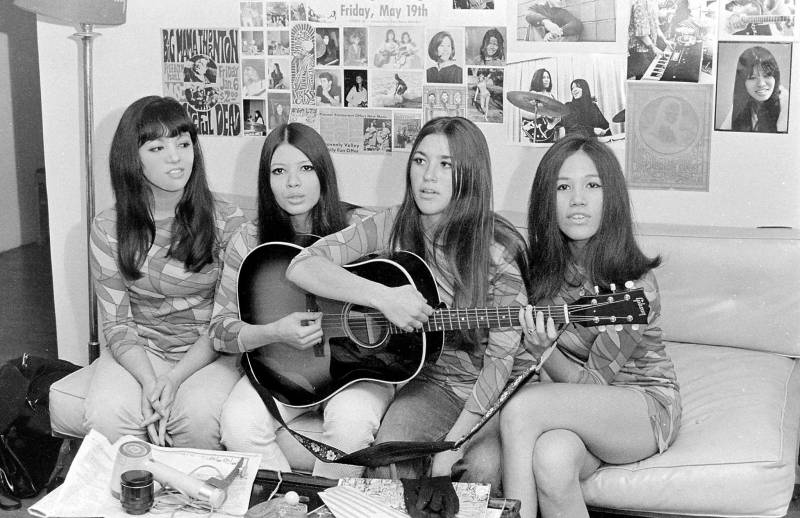 A vintage black and white photo of four women sitting on a couch with three of them holding a guitar with newspaper clips on the wall behind them.