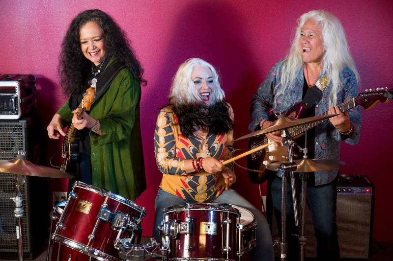 Three women playing a guitar, drums, and bass are seen in front of a pink-colored wall.