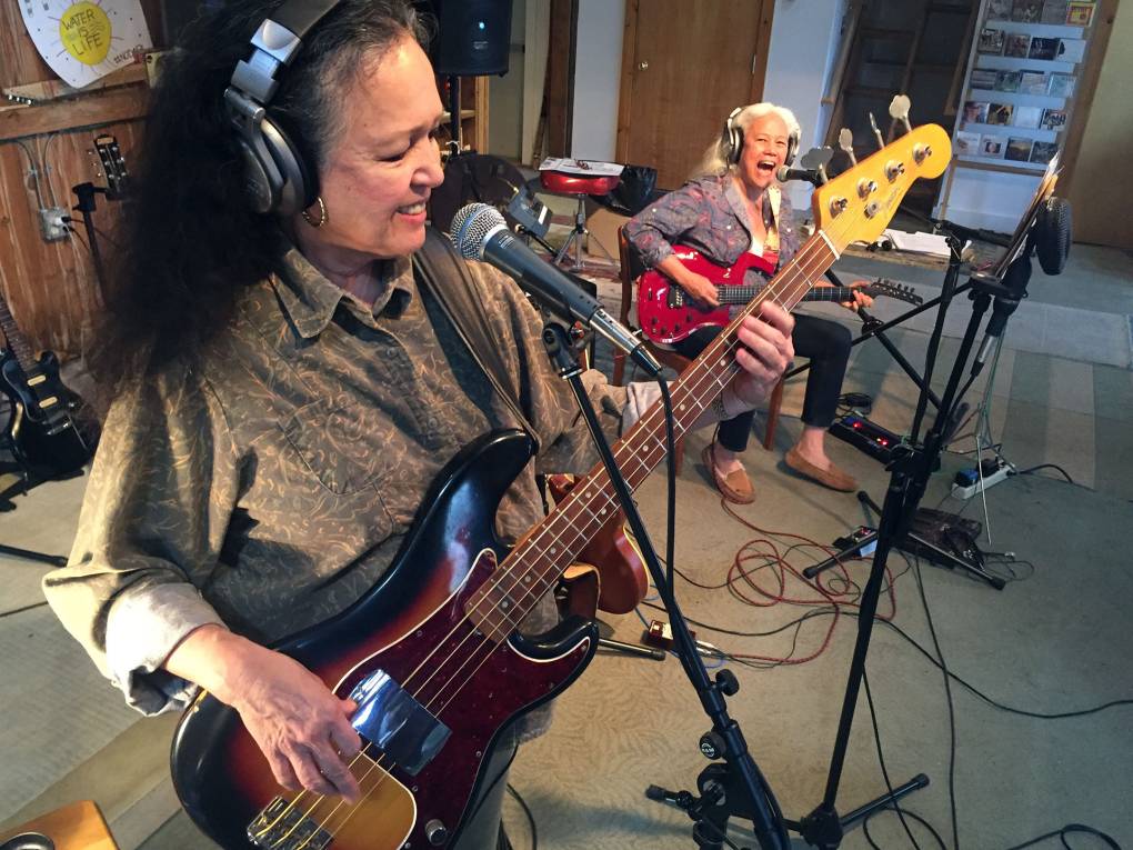 Two Filipina-American women in their late sixties rock out on bass and guitar in a recording studio, smiles on their faces.