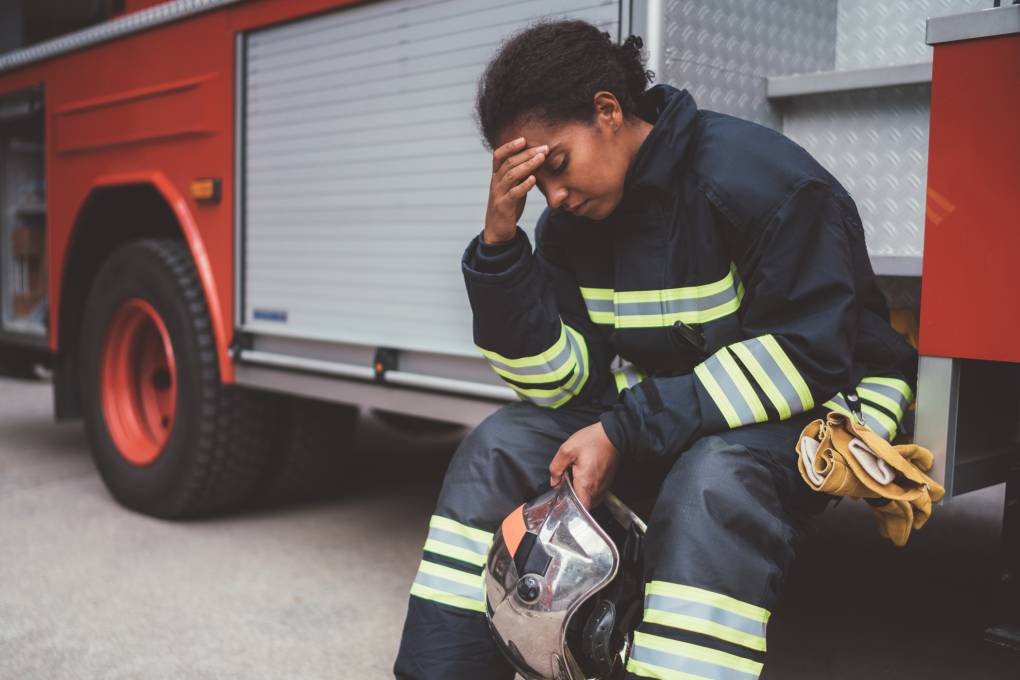 A woman wearing a firefighter uniform sits on the side of a fire truck with her hand massaging her forehead and the other hand holding a helmet.