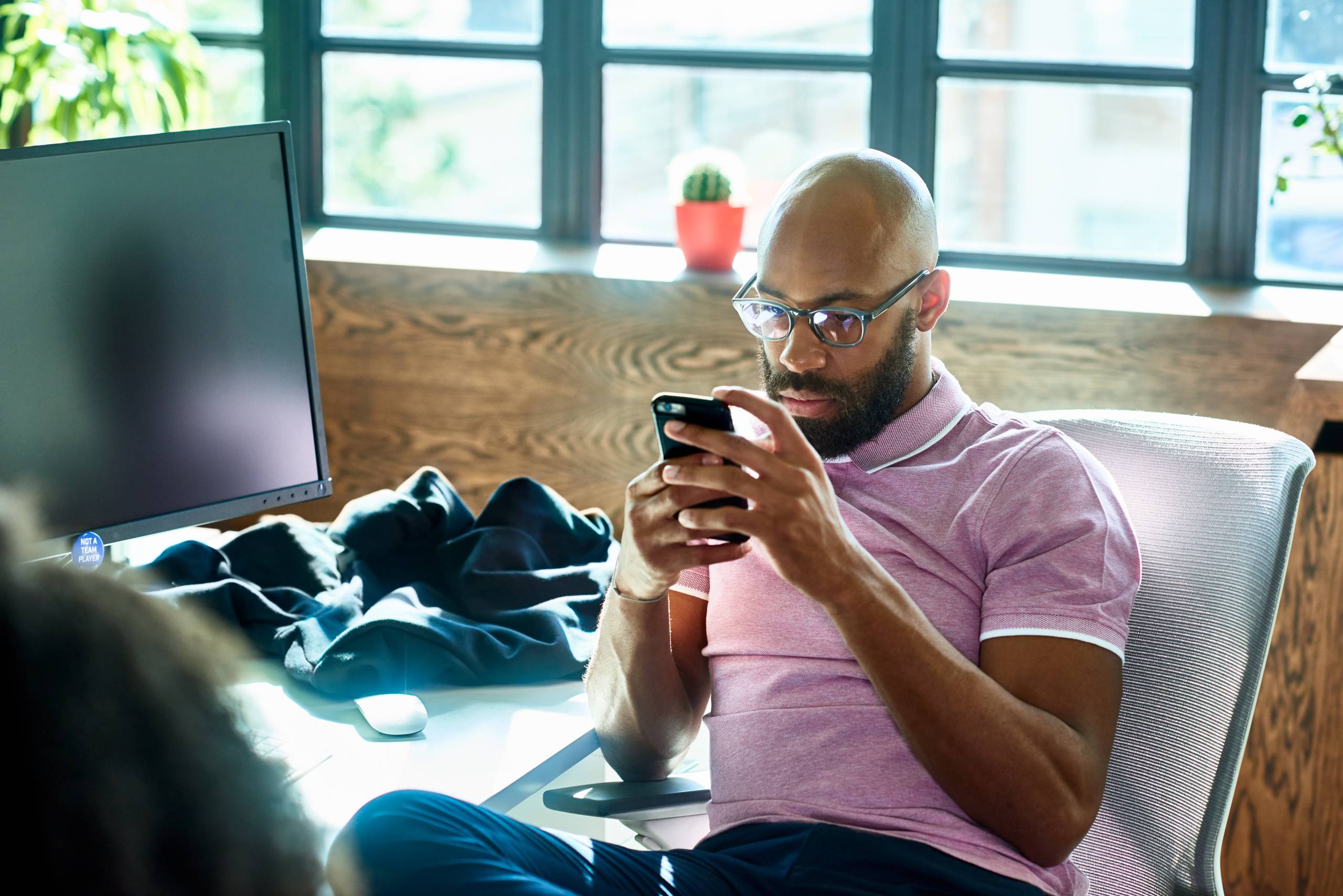 Man with beard and glasses looks at his phone screen while sitting next to a desk.