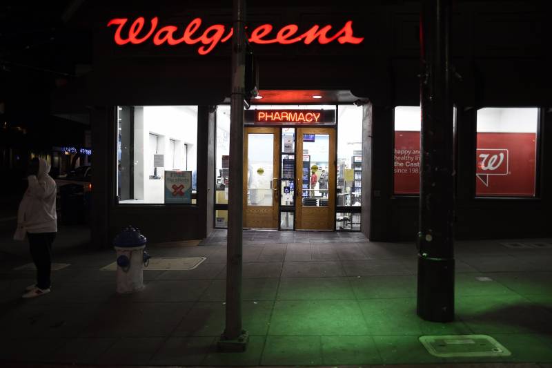 An after dark shot of the outside of the pharmacy, Walgreens. Its store name in cursive, red letters is illuminated above the entrance. A customer with a gray, hooded sweater stands near a fire hydrant outside of the store.