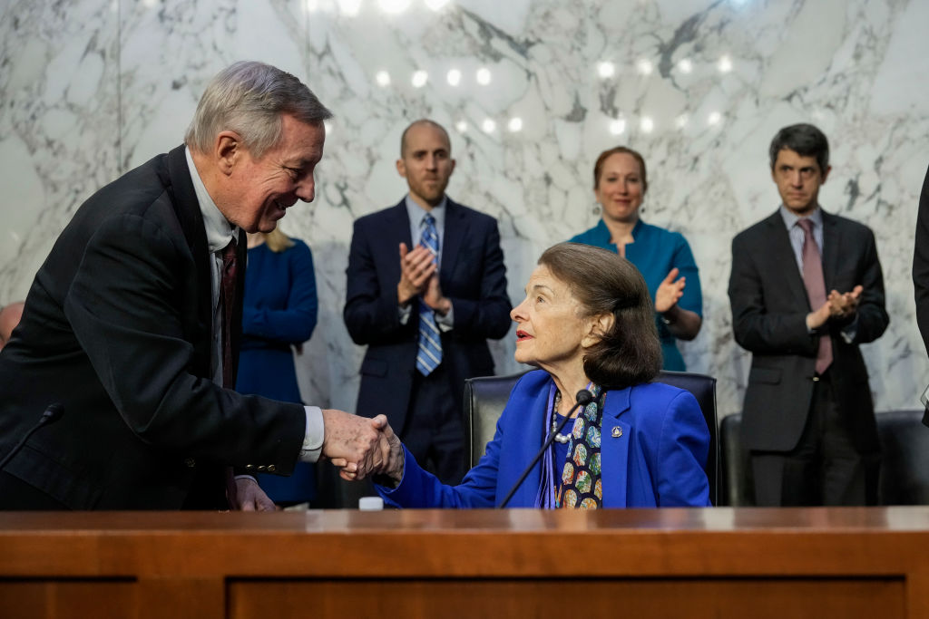 A seated older woman with a blue suit dress shakes hands with a smiling white man in a suit while others behind them clap their hands in a Senate judicial committee session.