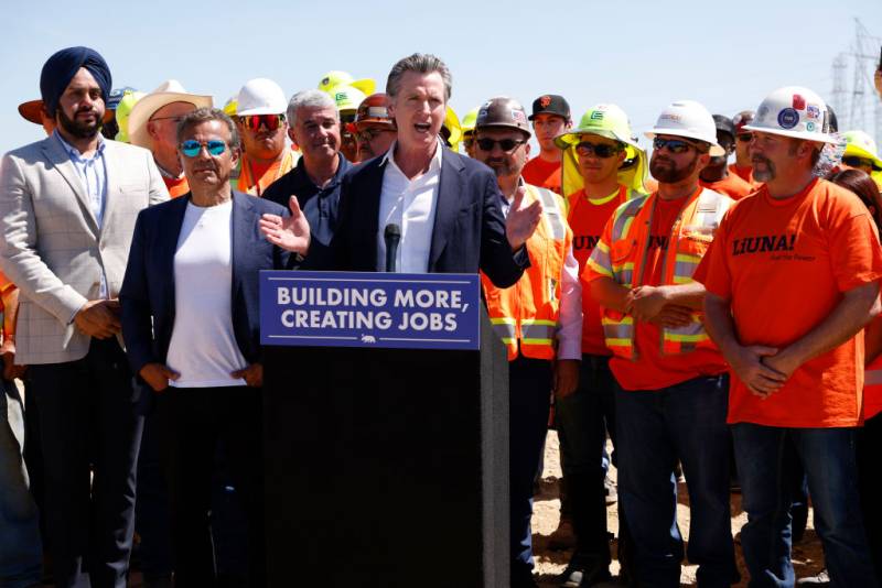 A white man talks to a crowd at a dais during a press conference with workers and others standing around him.