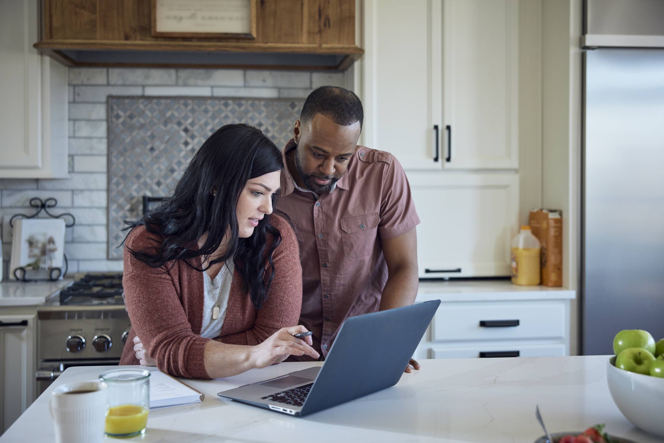 White woman and African American man sit at kitchen counter working together with a pen, paper and laptop.