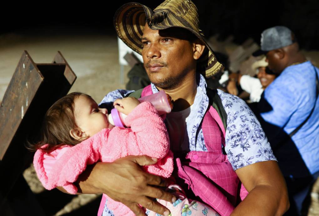A man wearing a hat and blue shirt holds a baby in a pink shirt with a bottle with other people in the background.