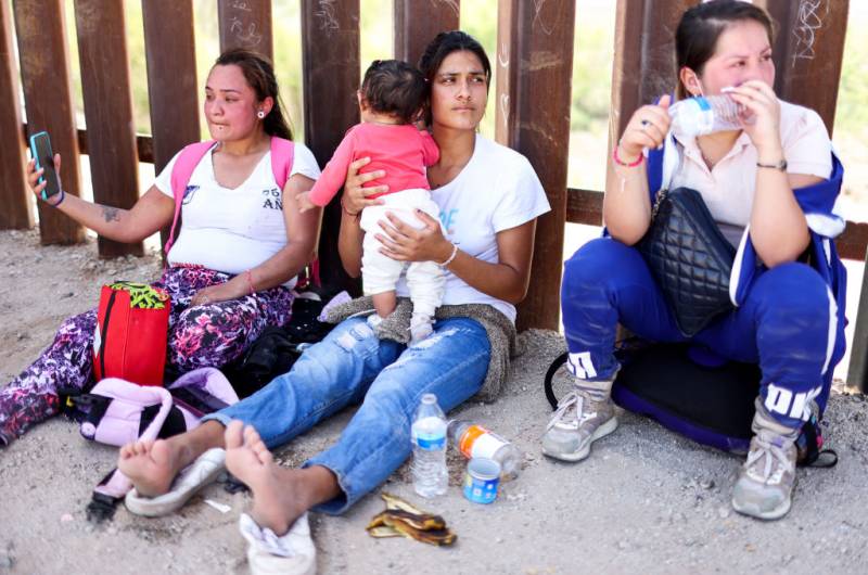 Three women wearing white shirts sit against a gate, with the woman in the middle holding a baby. Near them are water bottles and bags.