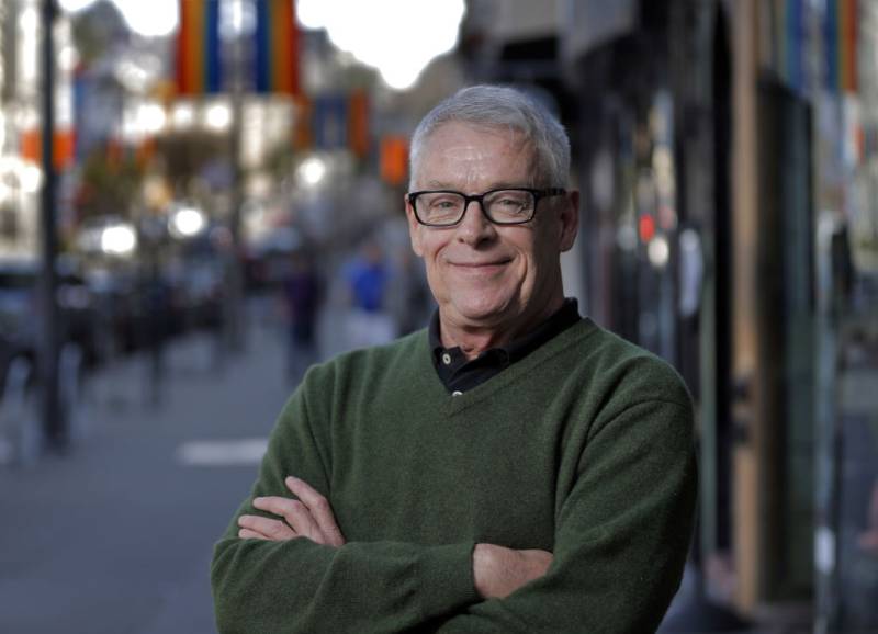 A bespectacled white man with white hair and a green sweater smiles at the camera with arms crossed and a blurry city street behind him.