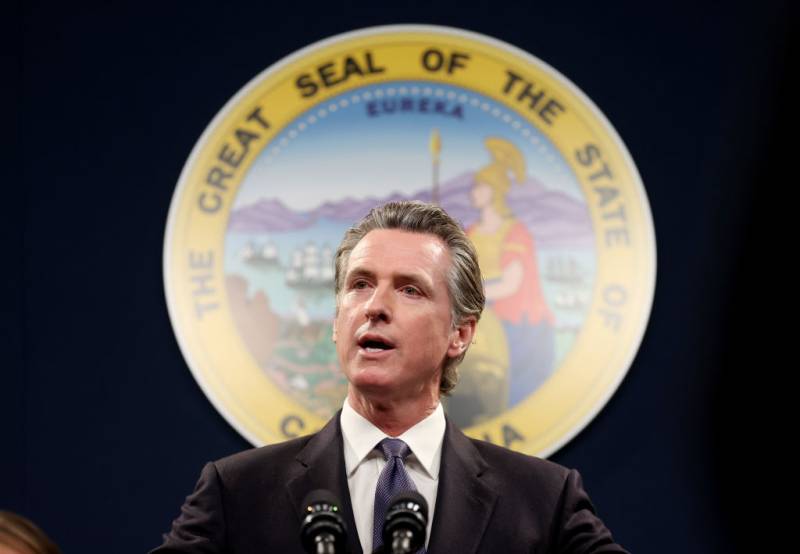 Headshot of a white middle-aged man speaking at a dais with the state seal of California behind him.