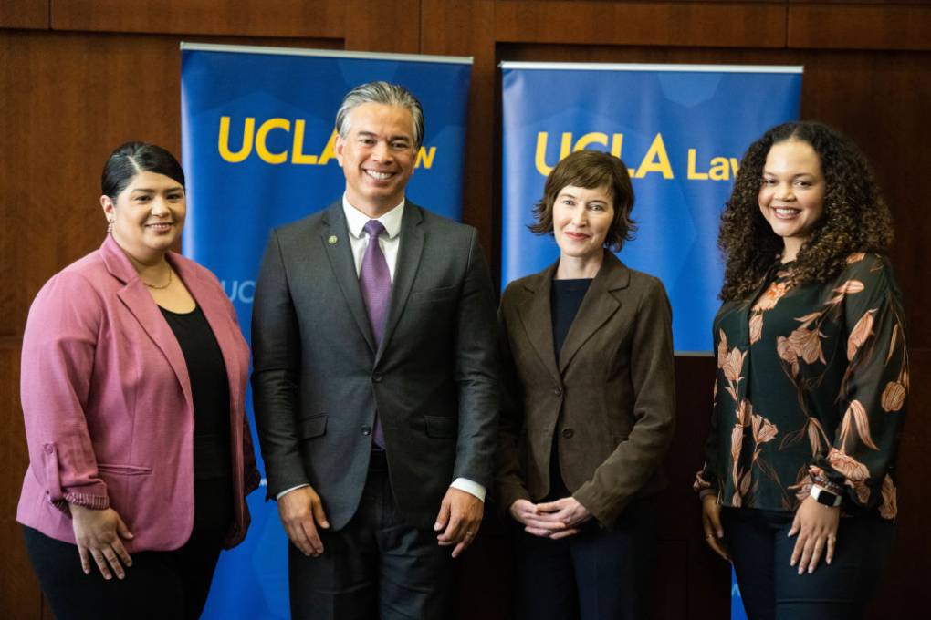 Three woman and a man stand and smile at the camera with a blue "UCLA Law" banner behind them.