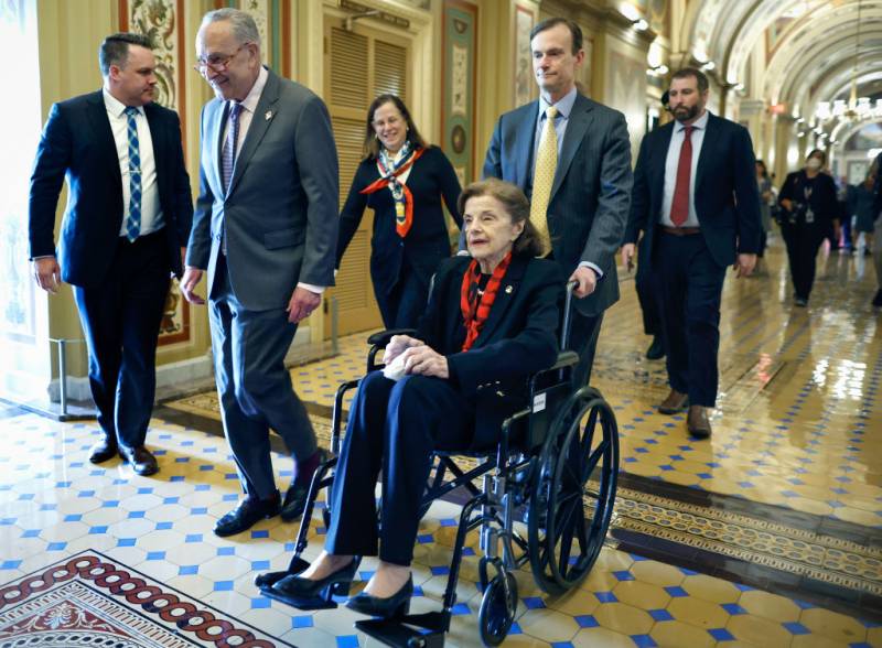 A white man pushes an older white woman in a wheelchair down a corridor in the US Capitol with others walking beside and behind them.