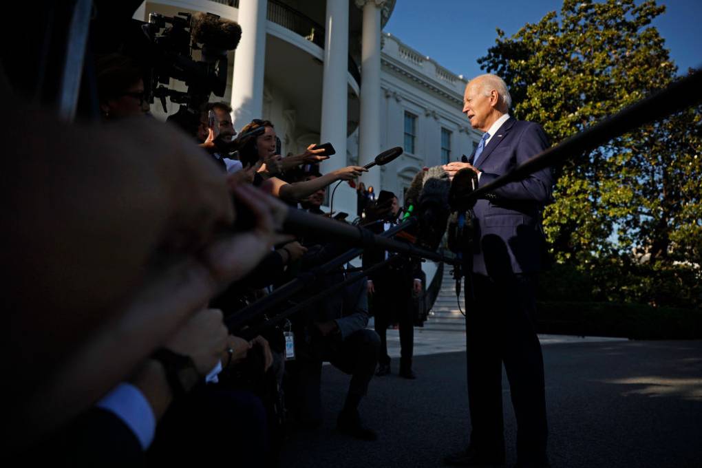 A white older man in a suit talks to reporters on a lawn outside a white building.