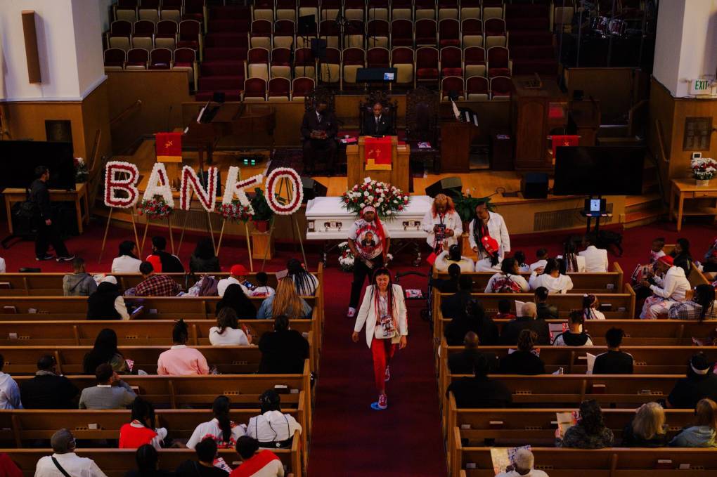 Inside a church the name "Banko" is spelled out in a floral display next to a white casket. Many sit in rows gathered to pay respect at this funeral.