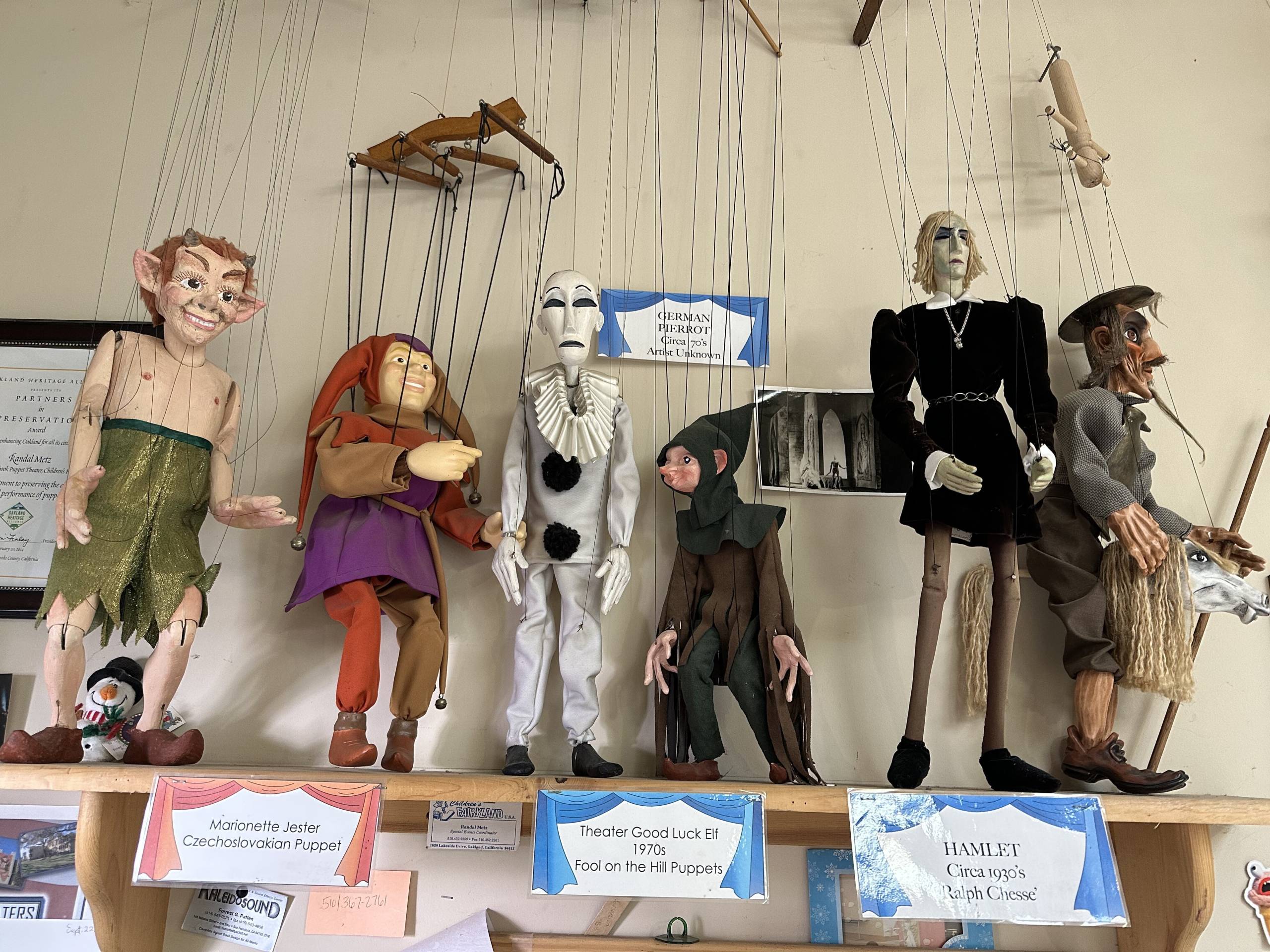 A view of six marionettes arranged on a high shelf, with varying styles, including a multicolored jester and a white-faced mime.