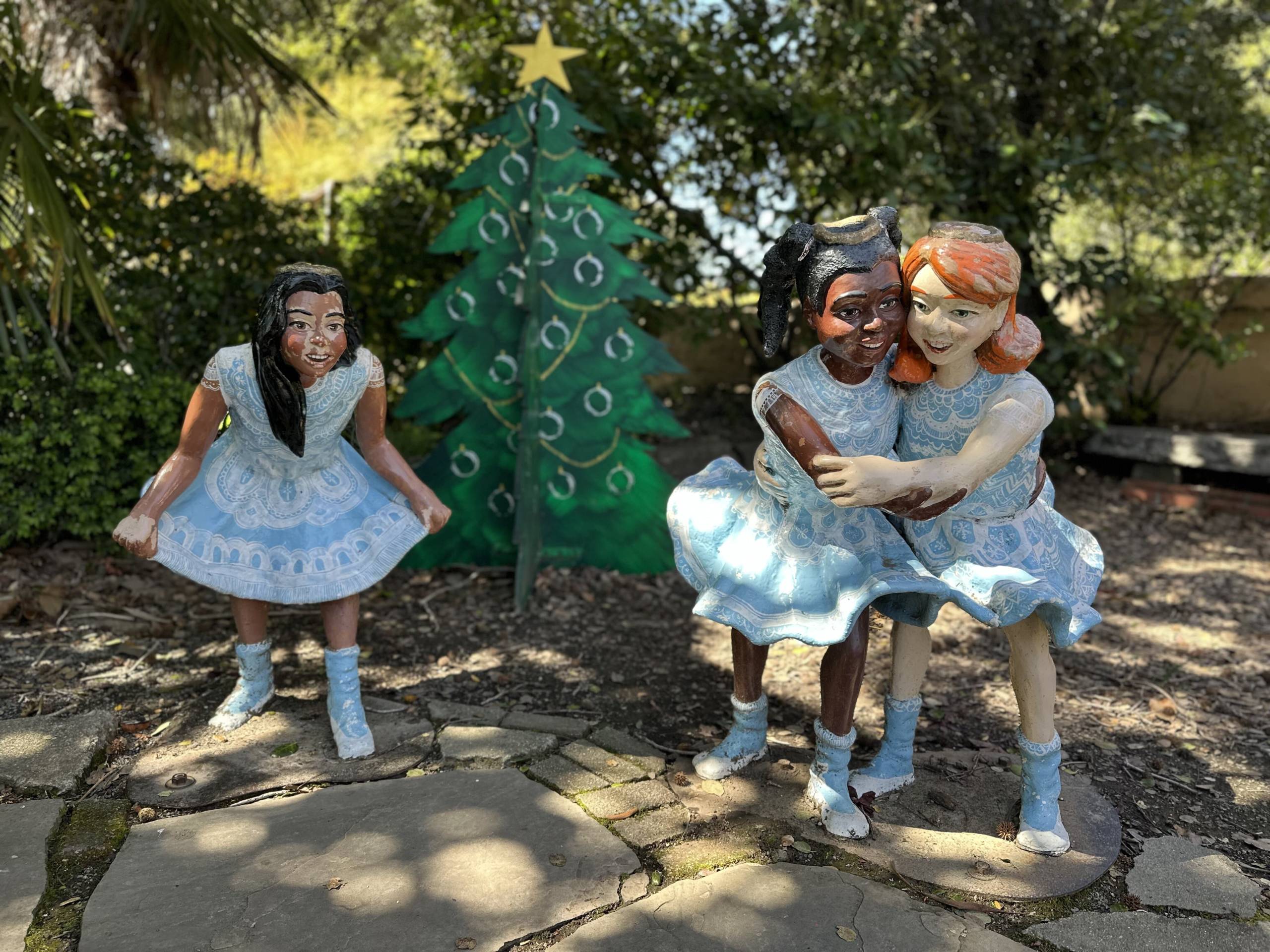 Three seemingly life-size statues of smiling little girls in white-and-blue pinafores. The girl on the far left appears Asian and has long black hair. The two girls to the right embrace happily; the girl on the left appears Black, with Black hair, and the third girl appears white, with red hair. They all stand in dappled sunlight beneath trees.