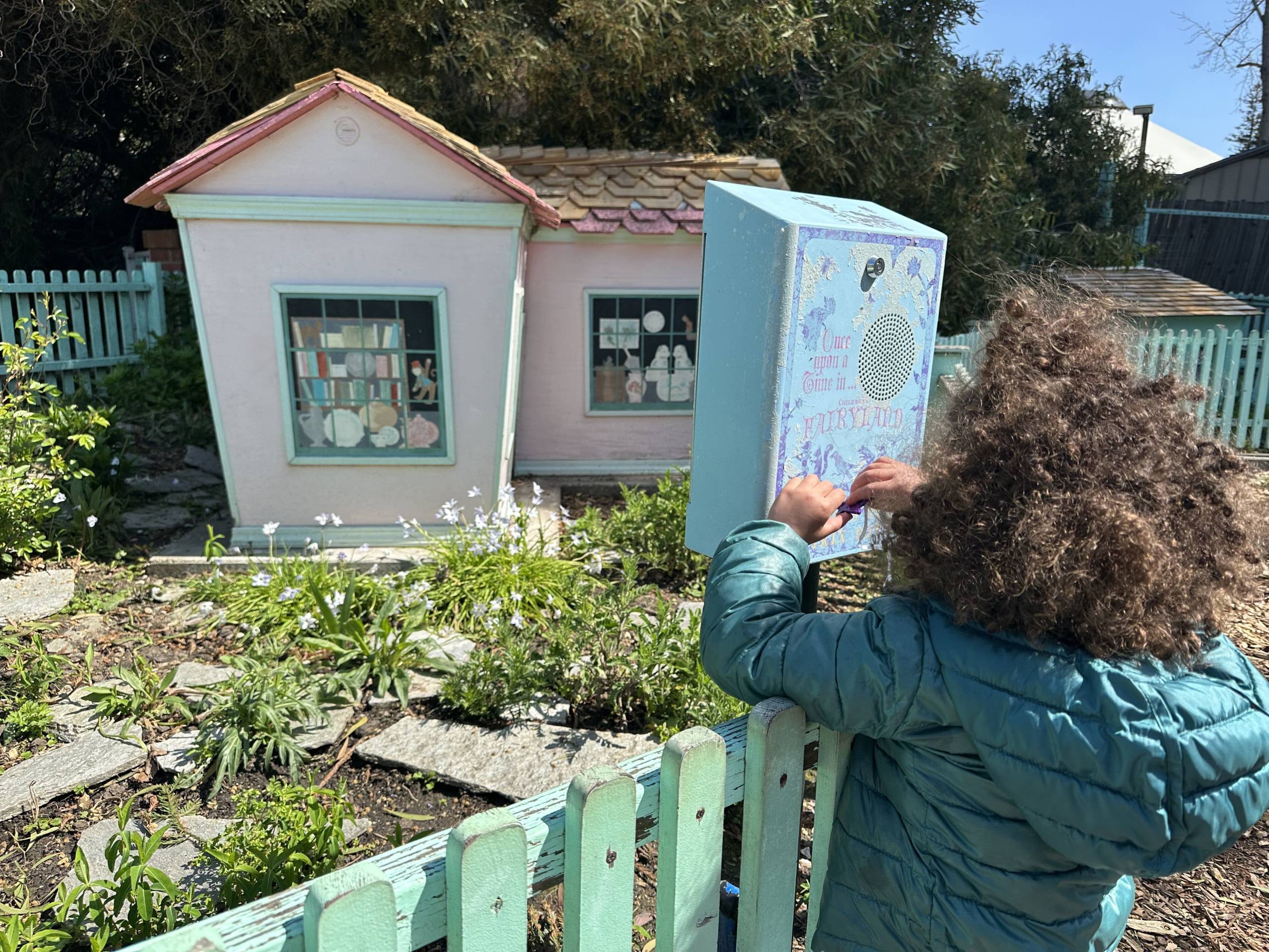 A little kid, around age 6, with poofy, coily hair and a blue puffer jacket turns a key in a light-blue wooden box. Beyond the box, which sits atop a turquoise-painted fence, is a little, light-pink house and flowers blooming amid paving stones.