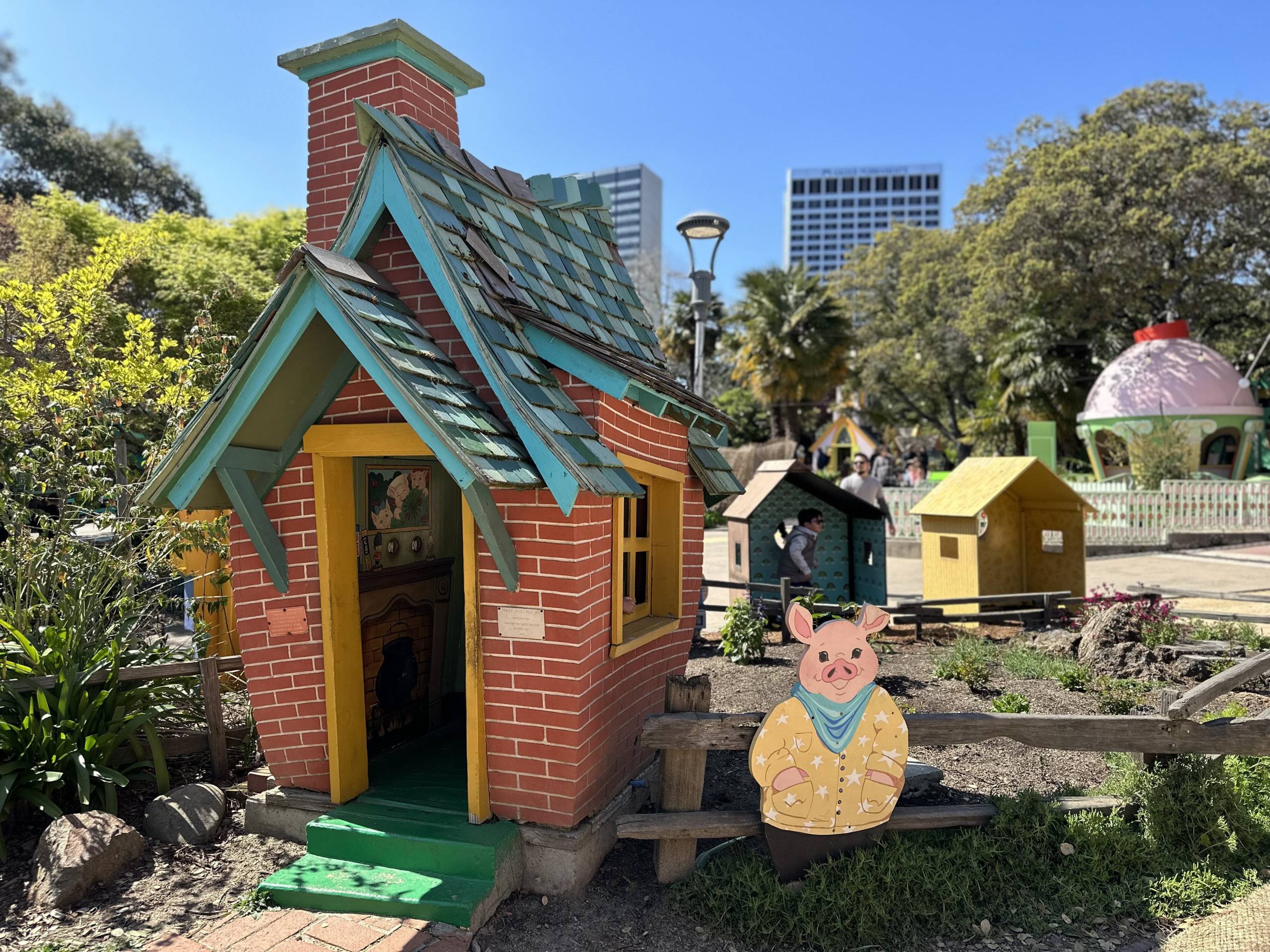 A three little piggies-themed play set, with a small brick house with a low doorway, and a cut-out cartoon pig next to it. A couple high-rise buildings in downtown Oakland are visible in the background.