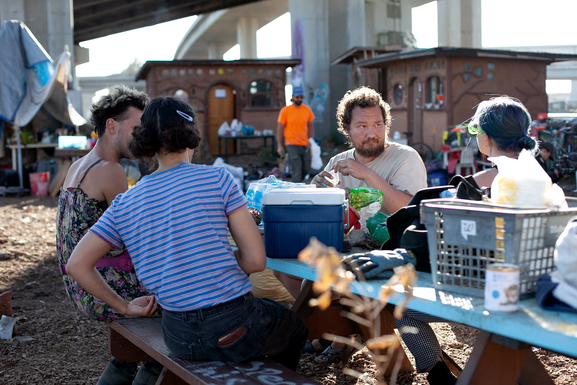 Four people sit at a picnic bench talking to one another. A small cooler sits on top of the table along with a gray basket. Tiny homes are pictured in the background that sit under a freeway overpass.