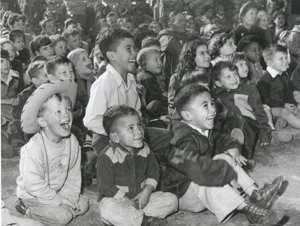 A black-and-white photo of dozens of children sitting on the ground, looking past the camera toward an unseen stage, and laughing really hard. The four boys in the foreground are dressed in cowboy gear, with Western shirts and one wearing a cowboy hat. Most of the children appear Latino and white.