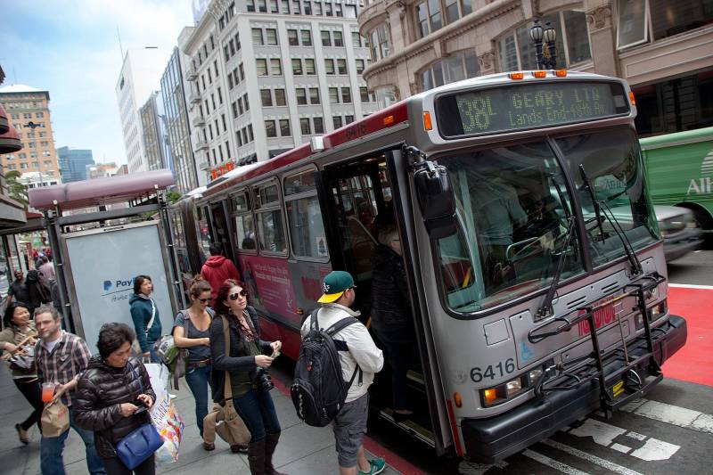Passengers board a bus on a busy street in San Francisco. One man is wearing an Oakland A's baseball cap.