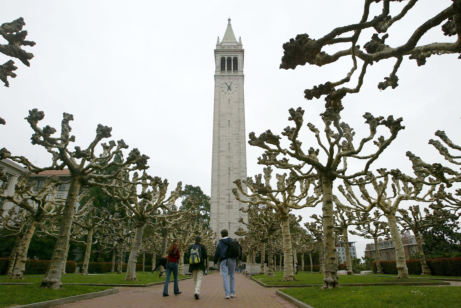 A tall, white clock tower punctuates the middle of a college campus outdoor space. Sections of grass and chunky trees are scattered throughout. The sky is gray. Three college students walk down a brick pathway together toward the clock tower. They wear book bags and jackets.