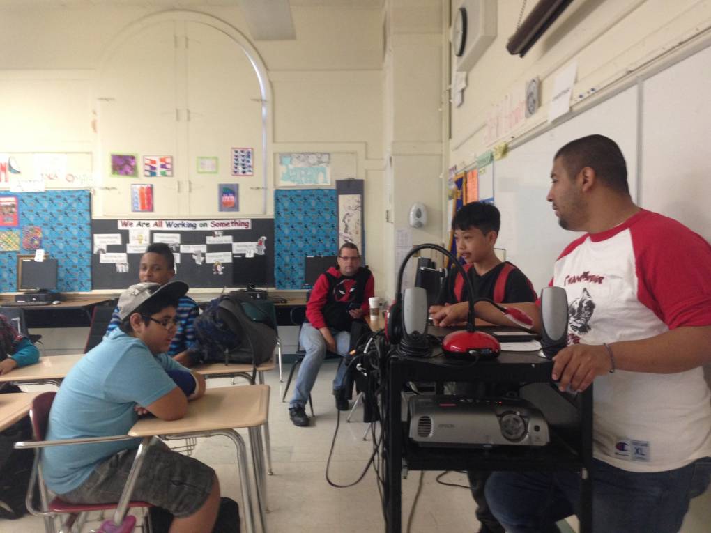Inside a classroom, students sit at desks as a teacher wearing a red and white T-shirt stands behind a projector as he talks to his students.