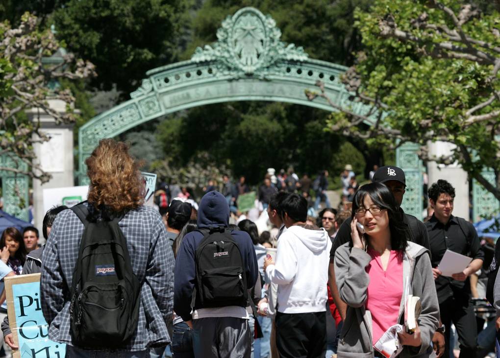 College students walk through a large, green archway surrounded by lush, tall trees. Many students wear black Jansport brand backpacks. Another female student with black hair and glasses talks on her cellphone in the foreground.