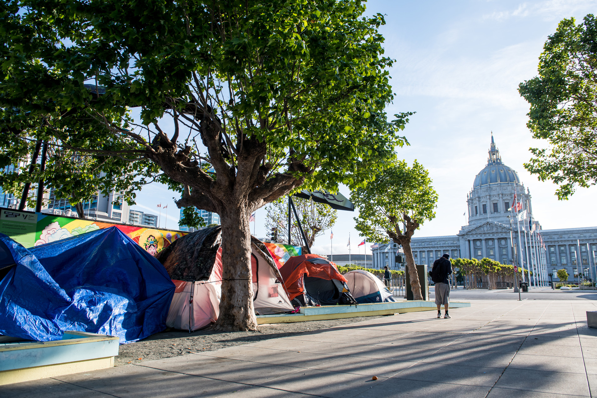 A row of tents line a sidewalk in downtown San Francisco near City Hall. The white, arched government building is seen standing tall in the background as the sun peaks over a few green trees. A blue tarp hangs over one tent.