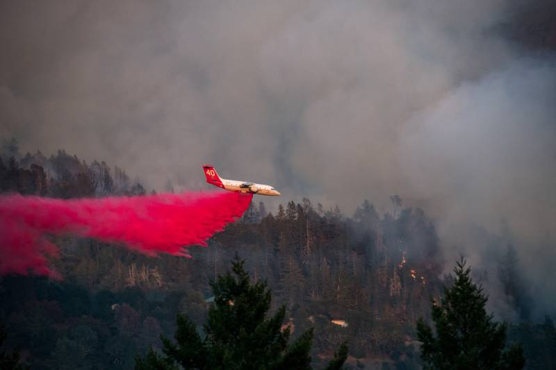 A white and red airplane is flying over hundreds of trees dropping red fire retardant spray all over the area to prevent a wildfire from spreading to nearby homes seen peeking through the trees below.