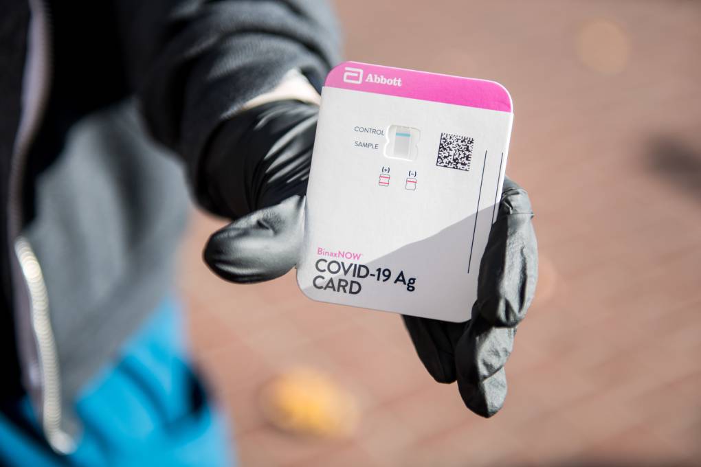 A hand wearing a black latex glove holds a COVID test card out to the camera