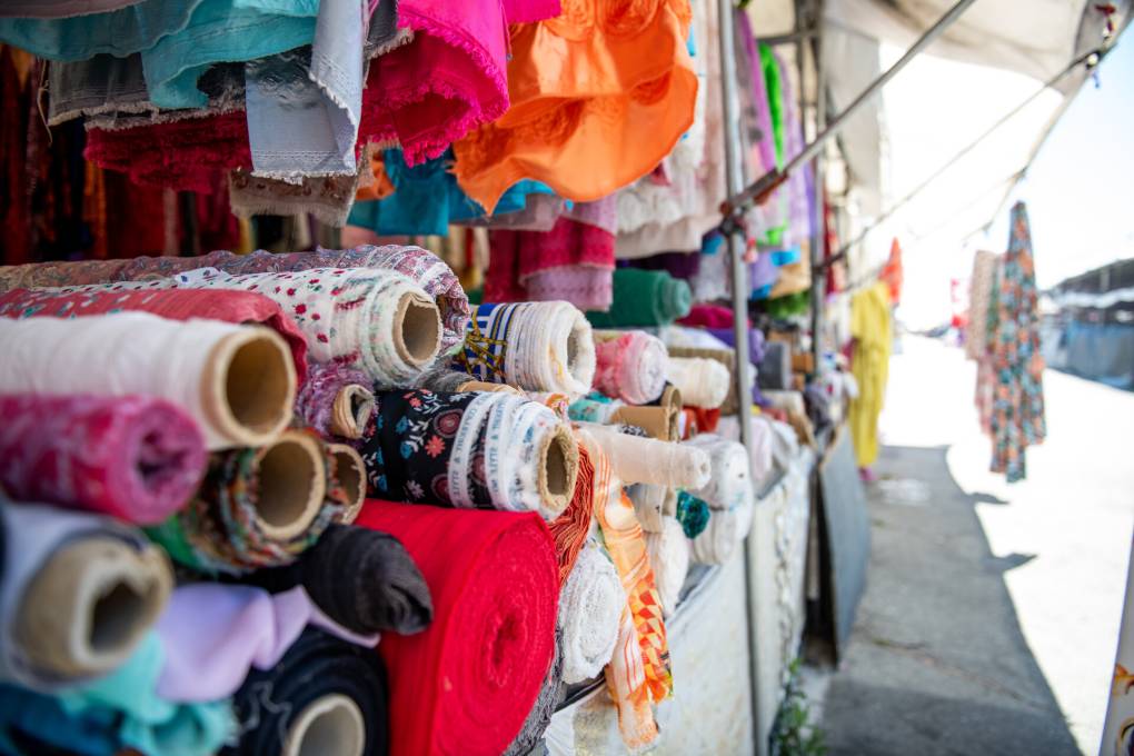 Large spools of fabric in various multicolor and floral patterns are piled on top of each other inside an outdoor vendor stall that sells at a local flea market. Garments are seen hanging in the background.
