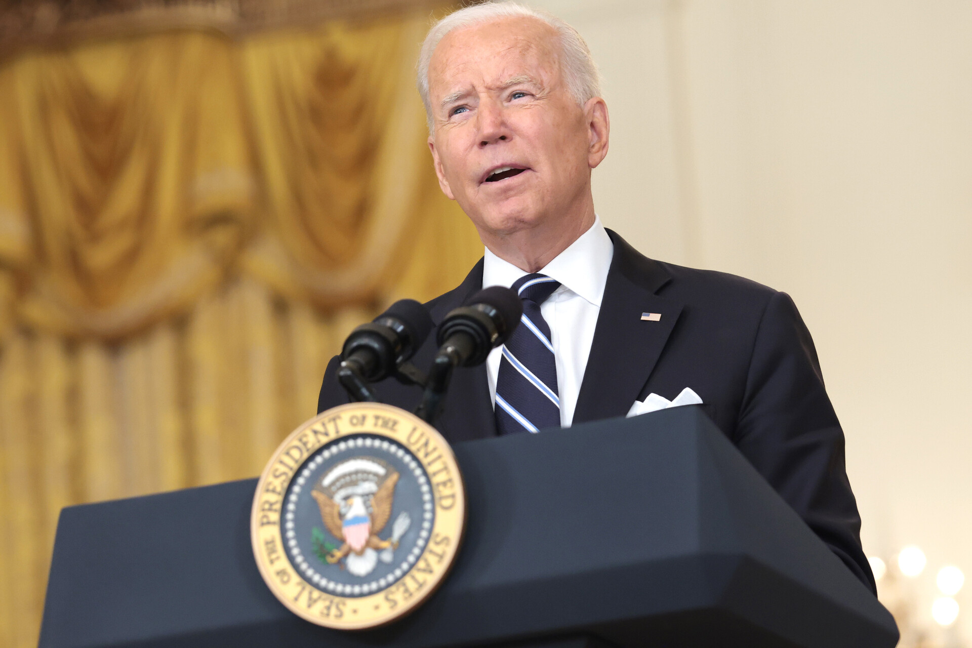 President Joe Biden speaks from the presidential podium inside the White House. He has short, gray hair, and wears a navy suit and tie. The presidential seal is in gold in the center of the podium and gold curtains are seen in the background.