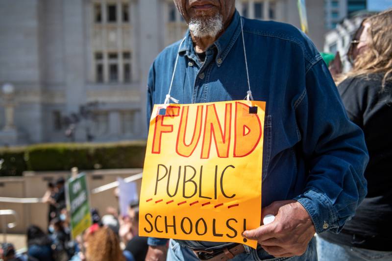 An older Black man stands outside at a rally, with a sign hanging from his neck that says: 'Fund Public Schools.'