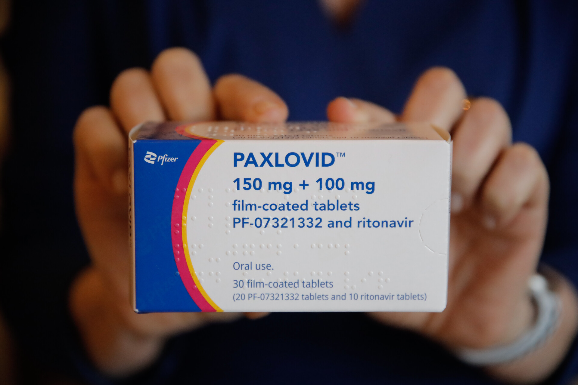 A white medication box with blue, pink and yellow stripes on the left-hand side. The words "Paxlovid 150 mg + 100 mg film-coated tablets" is printed in blue. A woman's hands are holding either side of the box. It's a close-up shot.