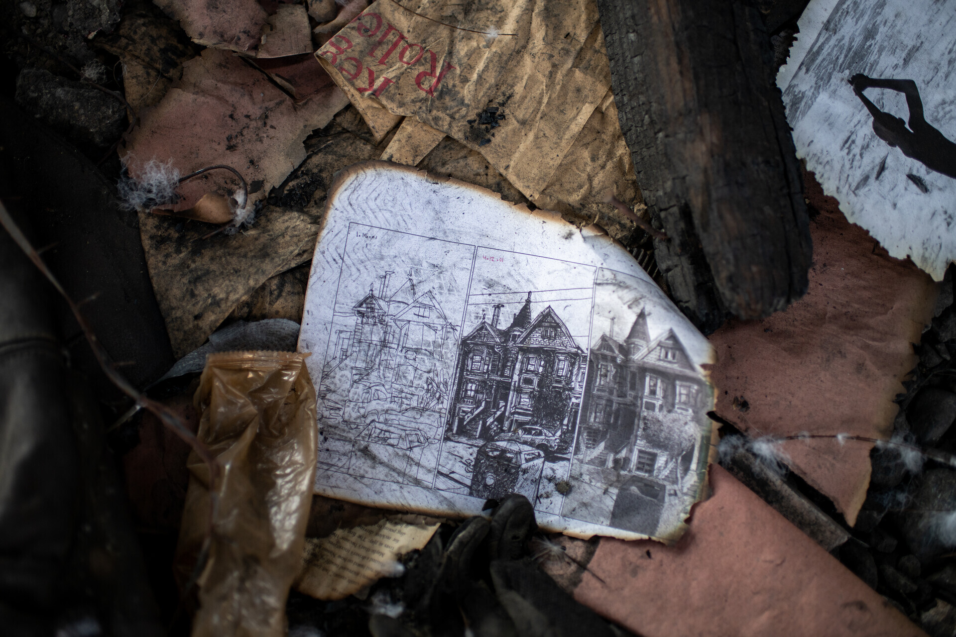 A white page with burned edges shows a charcoal drawing of Victorian facades, and sits among brown, shaded debris.
