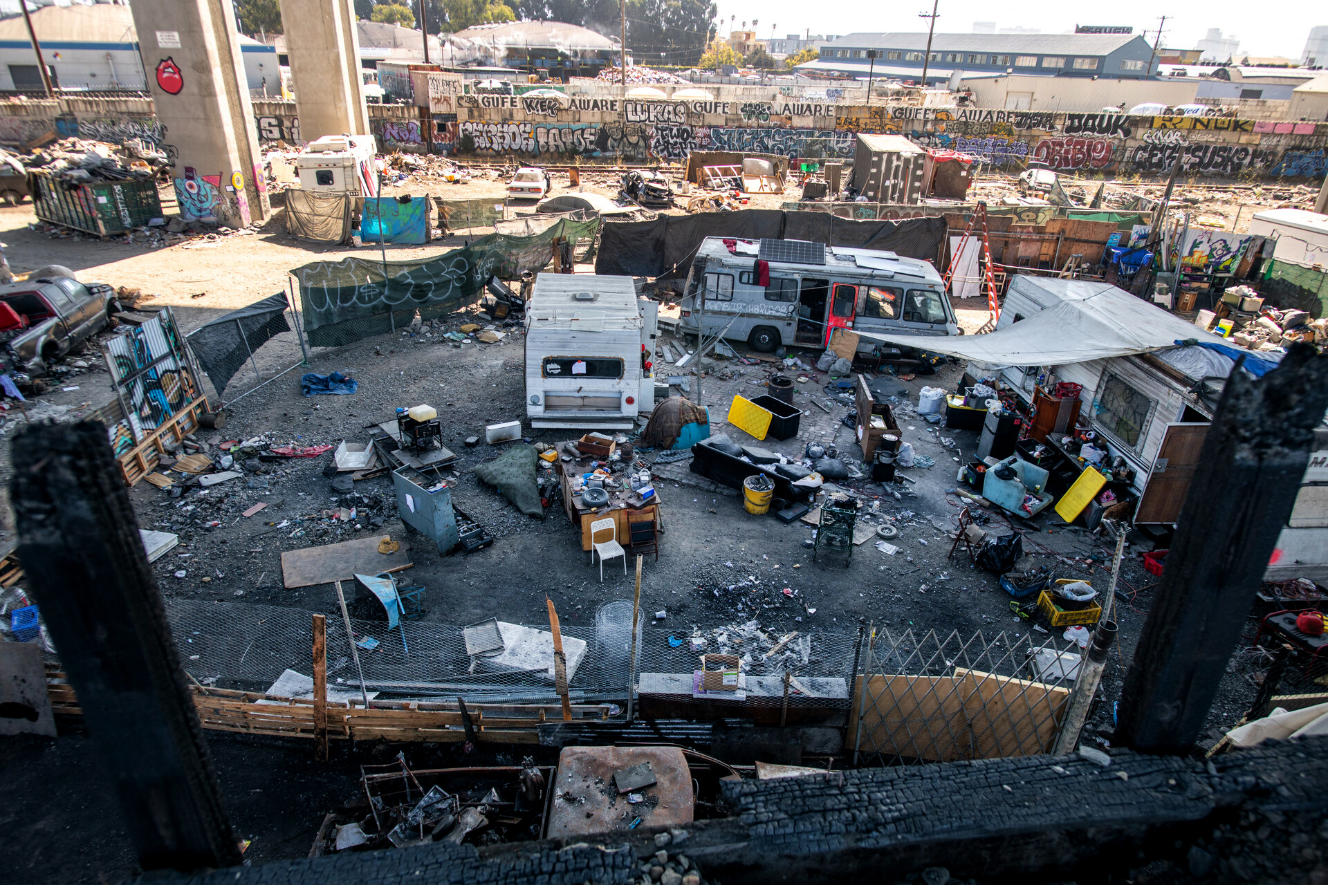 An aerial view of a homeless encampment with trailers, tents and people's belongings scattered about underneath a freeway overpass.
