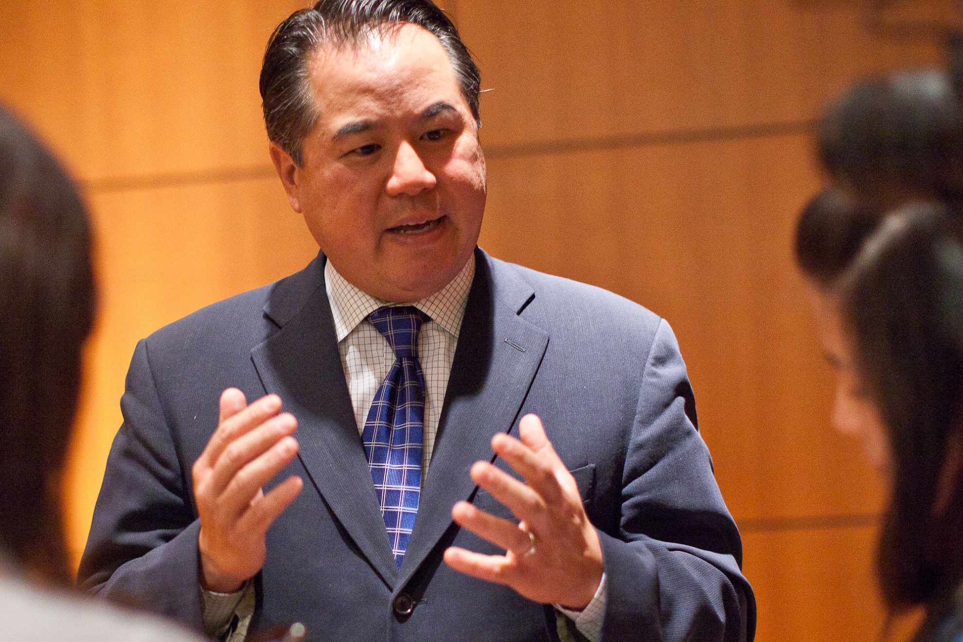 An Asian man with black hair combed back in a blue business suit is speaking with his hands.